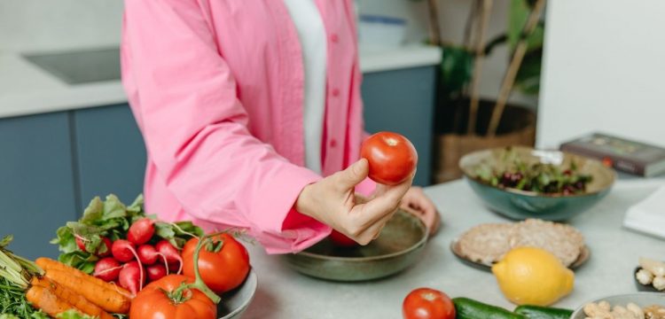 Person in pink overshirt holding a fresh tomato with various organic vegetables on a countertop.